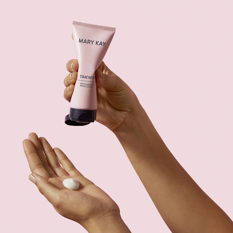 Mano apretando una cucharada de TimeWise Antioxidant Moisturizer en la otra mano contra un fondo rosa.